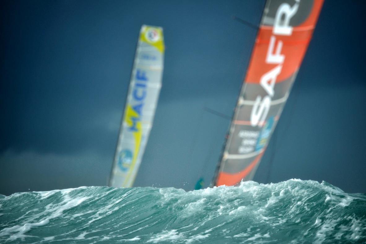 Voiles Macif et Safran émergeant d’une vague d’écume blanche sous un ciel sombre à Saint-Malo.