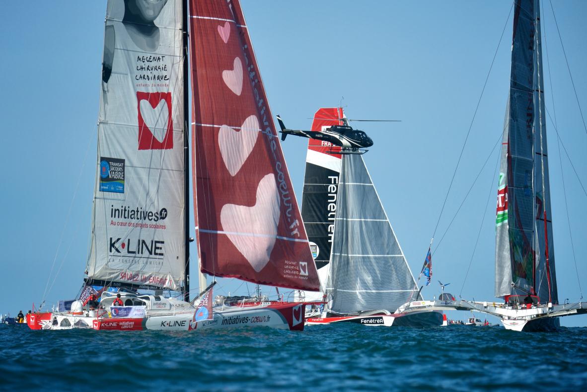Voiliers de course au large et hélicoptère de suivi sous un ciel bleu lors du départ de la régate au Havre.
