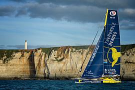 Voilier de course bleu devant les falaises de craie du Havre avec phare au sommet sous un ciel nuageux.