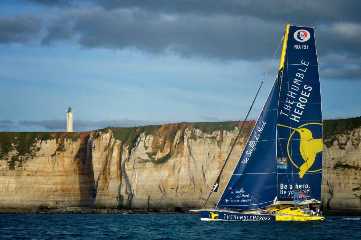 Voilier de course bleu devant les falaises de craie du Havre avec phare au sommet sous un ciel nuageux.