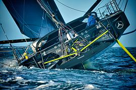 Voilier Imoca Hugo Boss noir gîtant sur l’eau bleue, foil jaune visible, skipper à la barre au Havre.