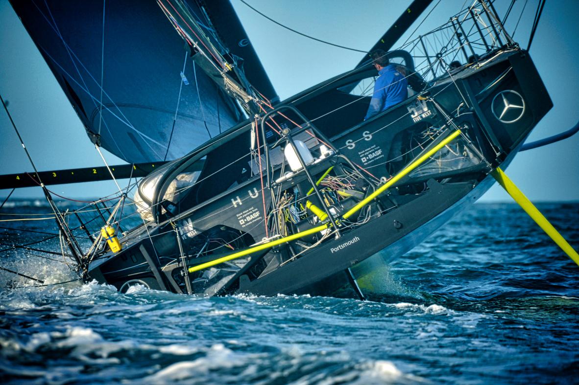 Voilier Imoca Hugo Boss noir gîtant sur l’eau bleue, foil jaune visible, skipper à la barre au Havre.
