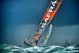 Voilier Imoca Safran penchant sur mer agitée, voiles oranges et noires, ciel sombre, Route du Rhum Saint-Malo
