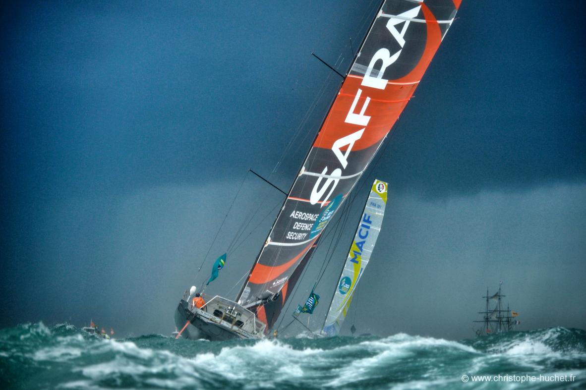 Voilier Imoca Safran penchant sur mer agitée, voiles oranges et noires, ciel sombre, Route du Rhum Saint-Malo