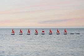 Huit optimists aux voiles rayées alignés sur une mer d’huile sous un ciel rose à Saint-Quay-Portrieux.