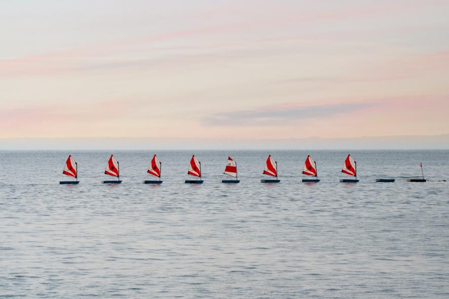 Huit optimists aux voiles rayées alignés sur une mer d’huile sous un ciel rose à Saint-Quay-Portrieux.
