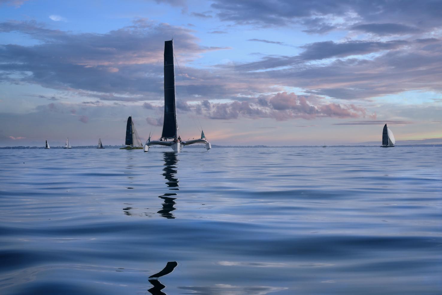 Plusieurs trimarans sur une mer calme aux reflets bleus sous un ciel nuageux de fin de journée en Bretagne.