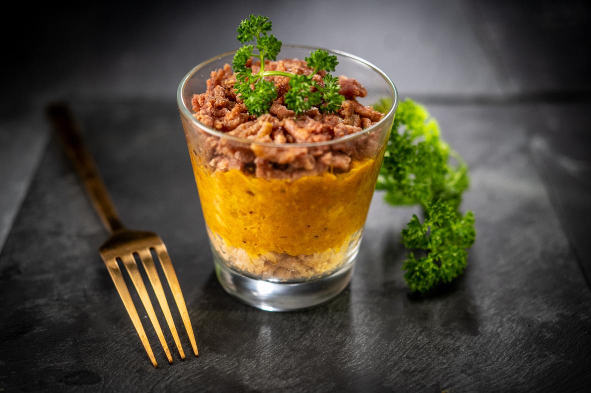 Verrine transparente aux couches de légumes et viande végétale sur ardoise noire avec fourchette dorée et persil frais.