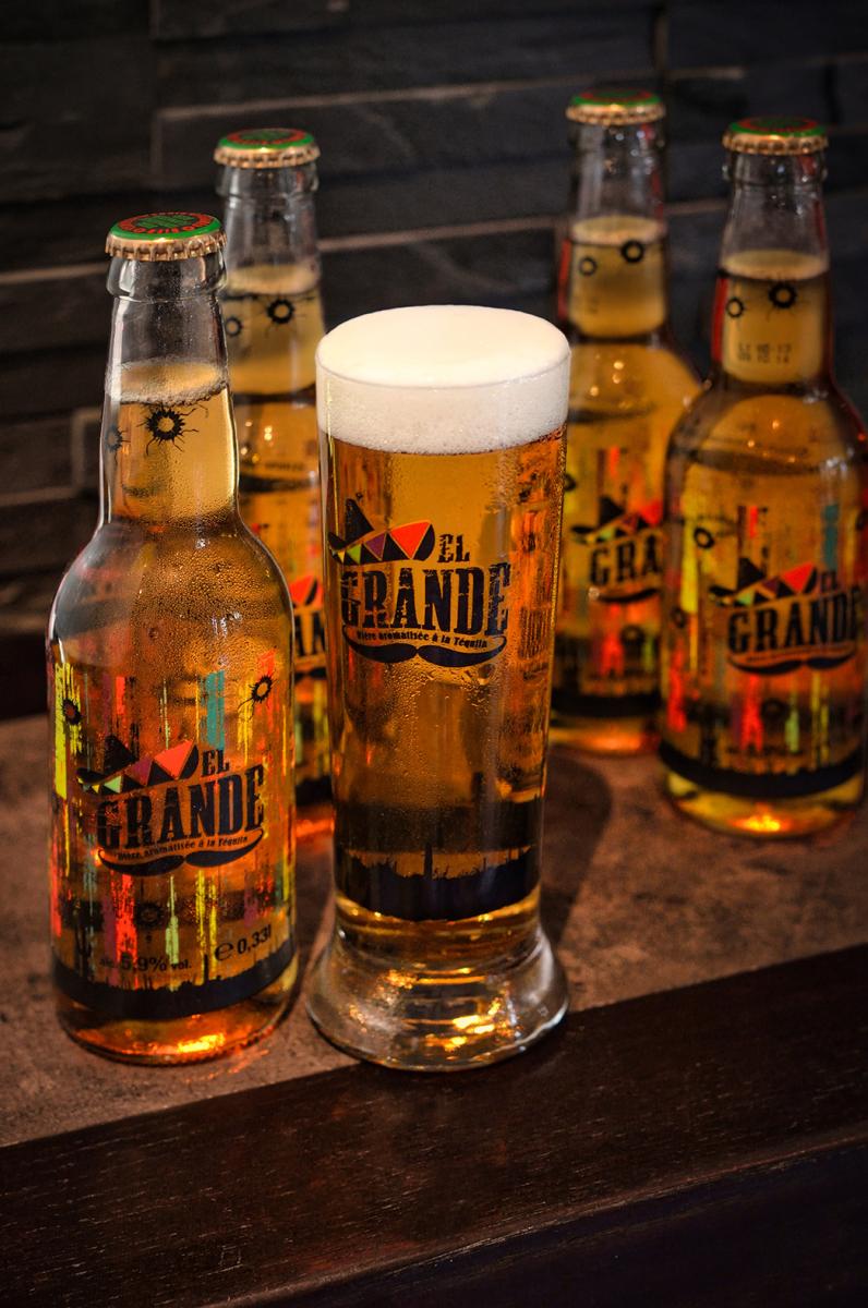Verre de bière blonde avec mousse épaisse devant quatre bouteilles de biere sur un comptoir sombre, éclairage chaleureu