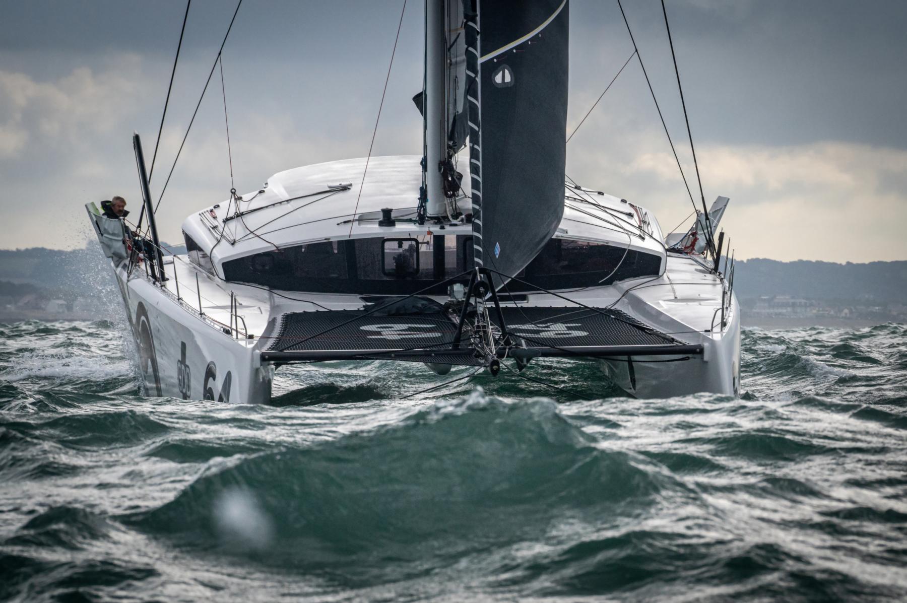 Catamaran de course GDD fendant les vagues, voiles sombres sur mer agitée par ciel nuageux, vue de face dynamique.