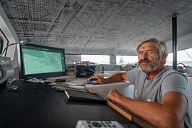 Portrait du skipper Halvard Mabire navigant sur son catamaran devant un écran de contrôle météo lumineux.