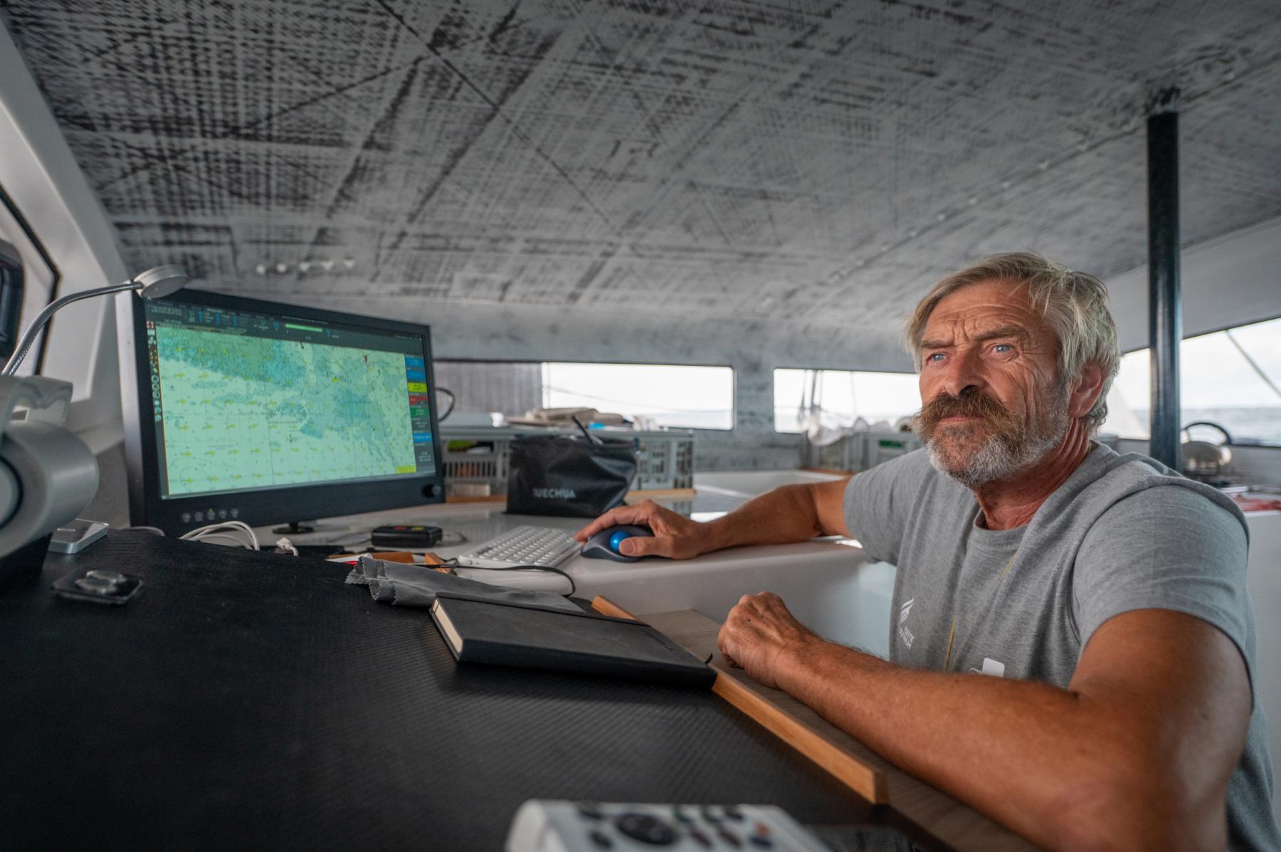 Portrait du skipper Halvard Mabire navigant sur son catamaran devant un écran de contrôle météo lumineux.