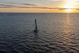 Vue aérienne du catamaran GDD naviguant sur l’océan au coucher du soleil, reflets dorés sur l’eau et horizon dégagé.