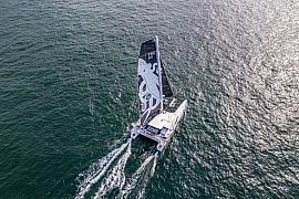 Vue aérienne d’un catamaran blanc sur mer scintillante avec voile noire illustrée d’un poulpe géant en pleine mer.
