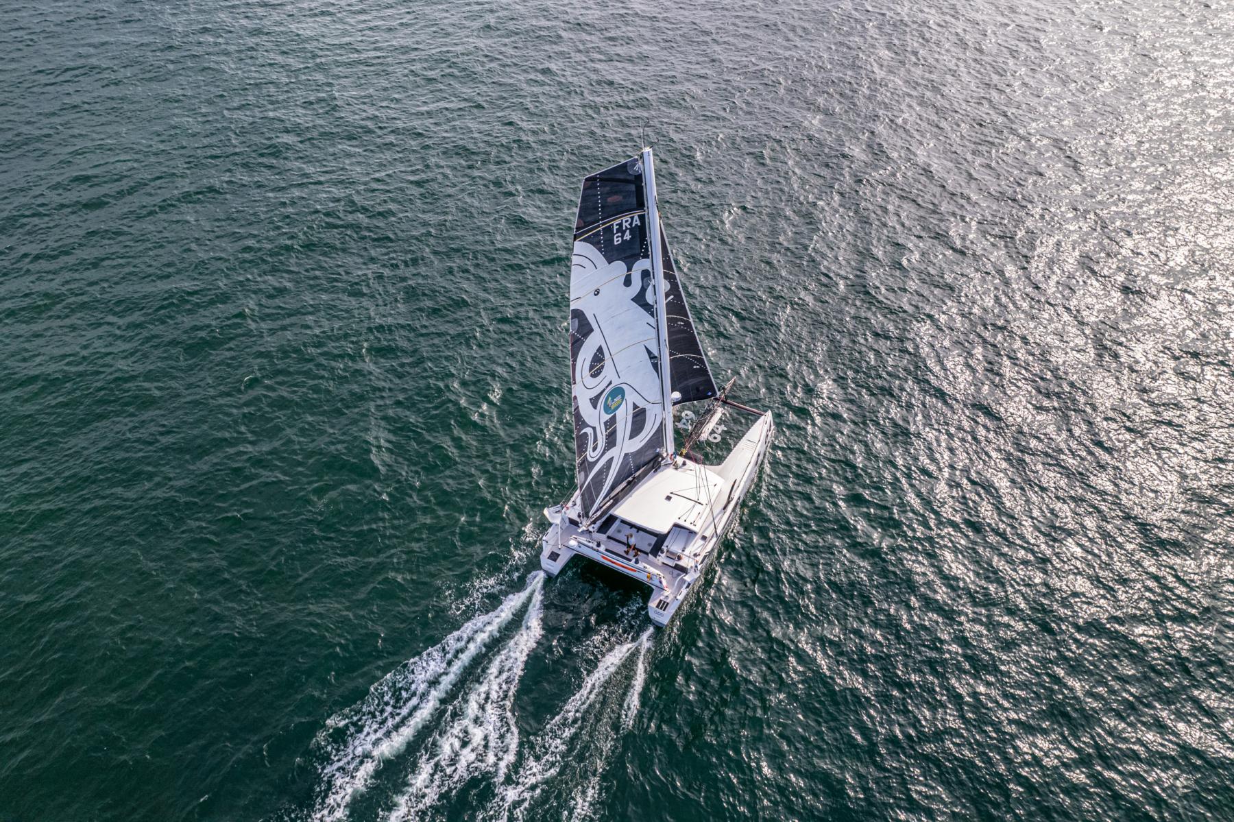 Vue aérienne d’un catamaran blanc sur mer scintillante avec voile noire illustrée d’un poulpe géant en pleine mer.