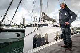 Skipper Halvard Mabire debout sur un quai devant le catamaran blanc GDD sous un ciel nuageux.