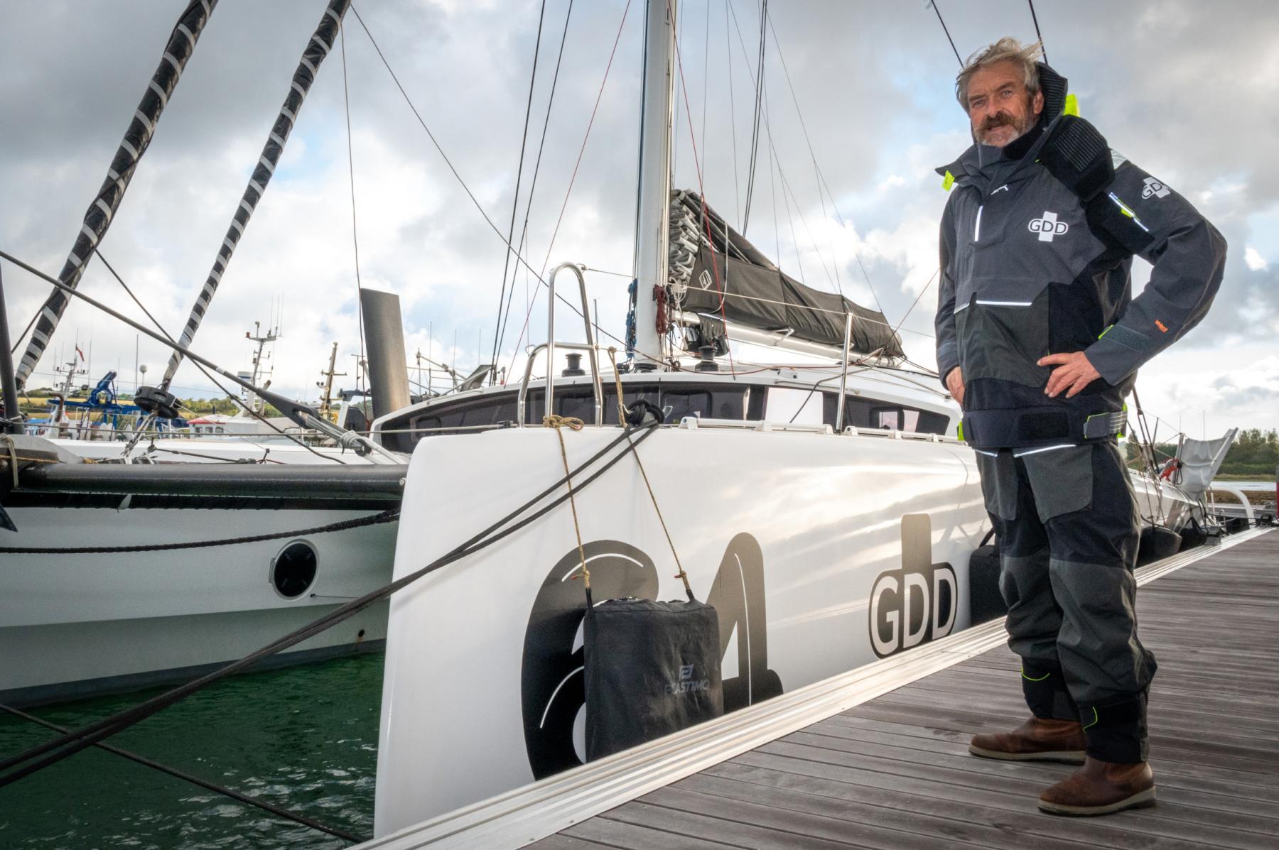Skipper Halvard Mabire debout sur un quai devant le catamaran blanc GDD sous un ciel nuageux.