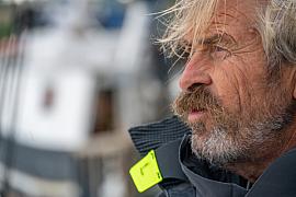 Portrait serré d’un skipper barbu aux yeux clairs en veste de quart technique sur un voilier, lumière naturelle de mer.