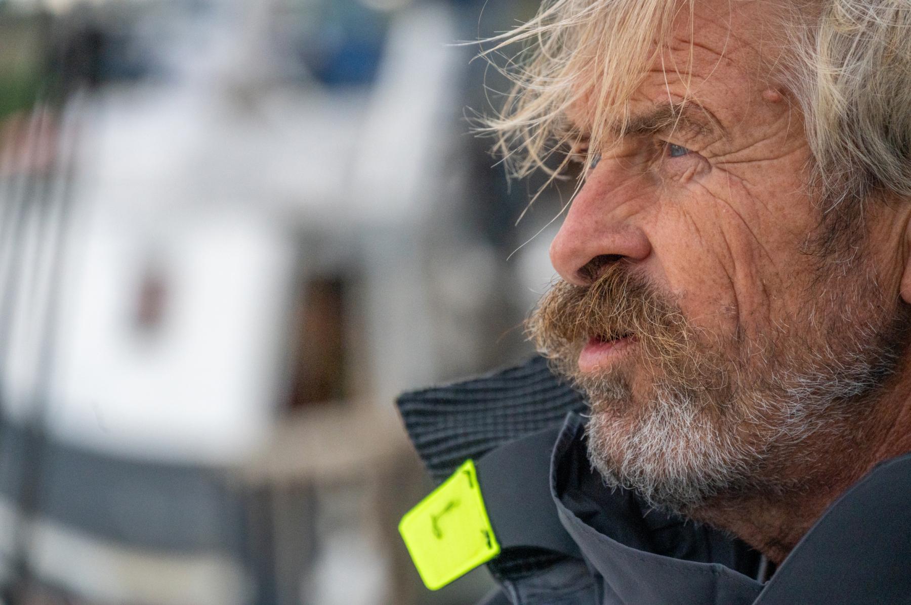 Portrait serré d’un skipper barbu aux yeux clairs en veste de quart technique sur un voilier, lumière naturelle de mer.