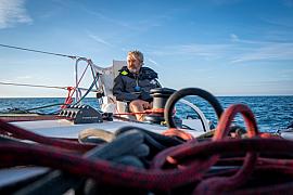 Skipper Halvard Mabire assis sur son catamaran GDD, cordages et bouts rouges au premier plan, mer bleue sous un ciel clair.