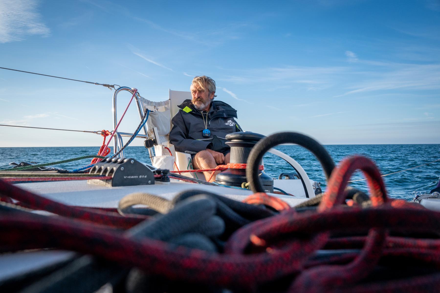 Skipper Halvard Mabire assis sur son catamaran GDD, cordages et bouts rouges au premier plan, mer bleue sous un ciel clair.