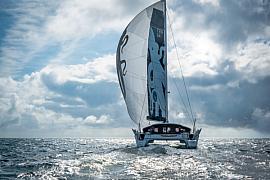 Catamaran GDD sous spinnaker blanc et noir naviguant en pleine mer sous un ciel nuageux rétro-éclairé.