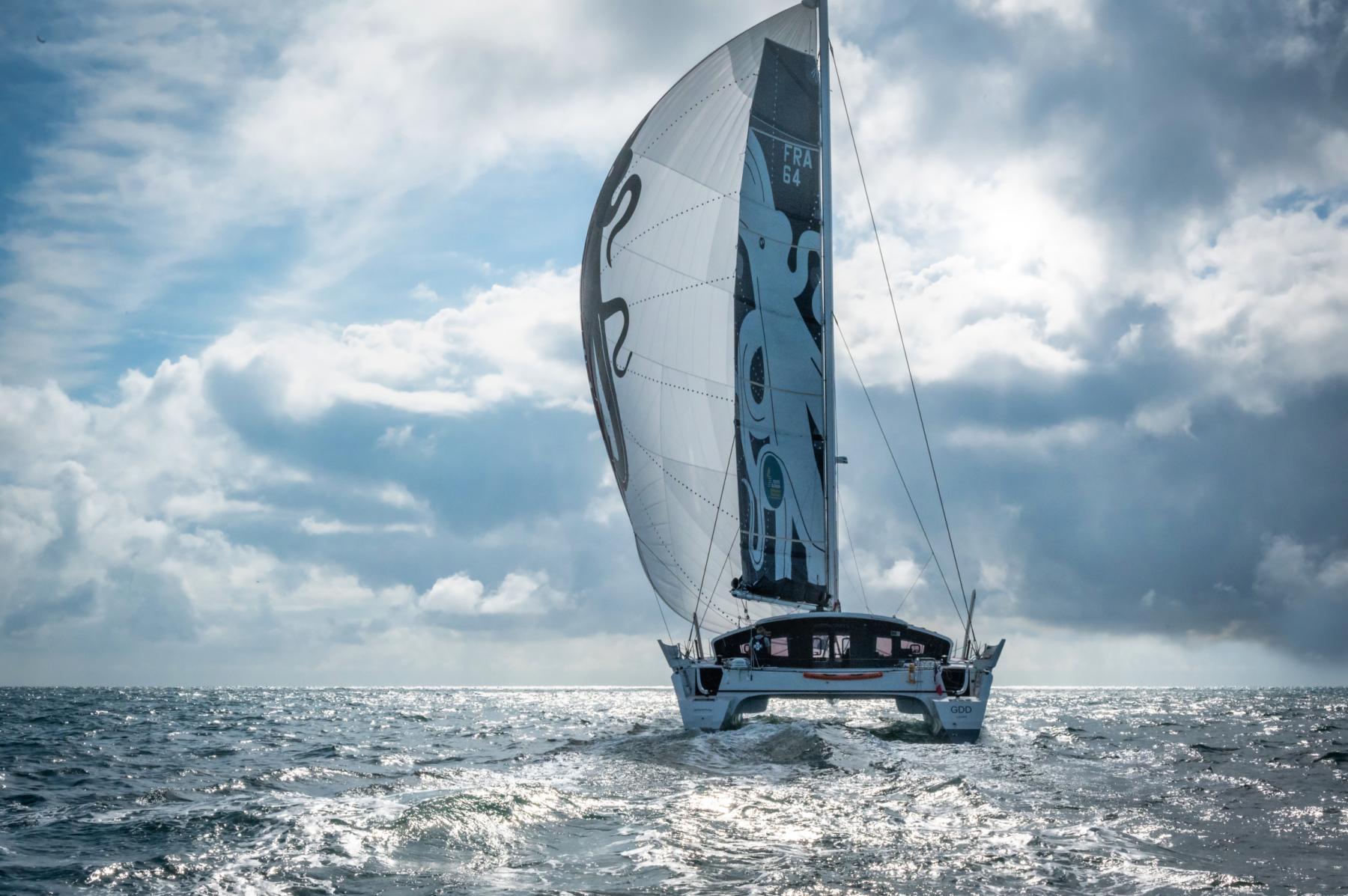 Catamaran GDD sous spinnaker blanc et noir naviguant en pleine mer sous un ciel nuageux rétro-éclairé.