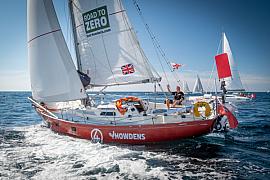 Voilier rouge Simon Curwen toutes voiles dehors sur mer bleue, skipper saluant de la main sous un ciel clair.