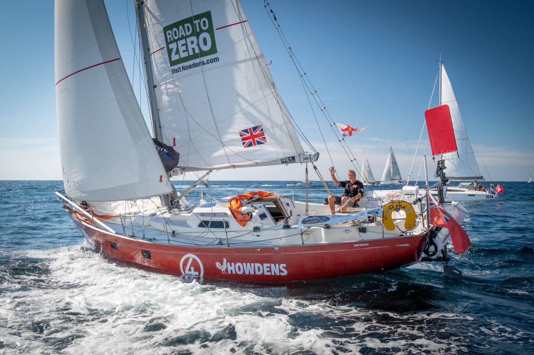 Voilier rouge Simon Curwen toutes voiles dehors sur mer bleue, skipper saluant de la main sous un ciel clair.