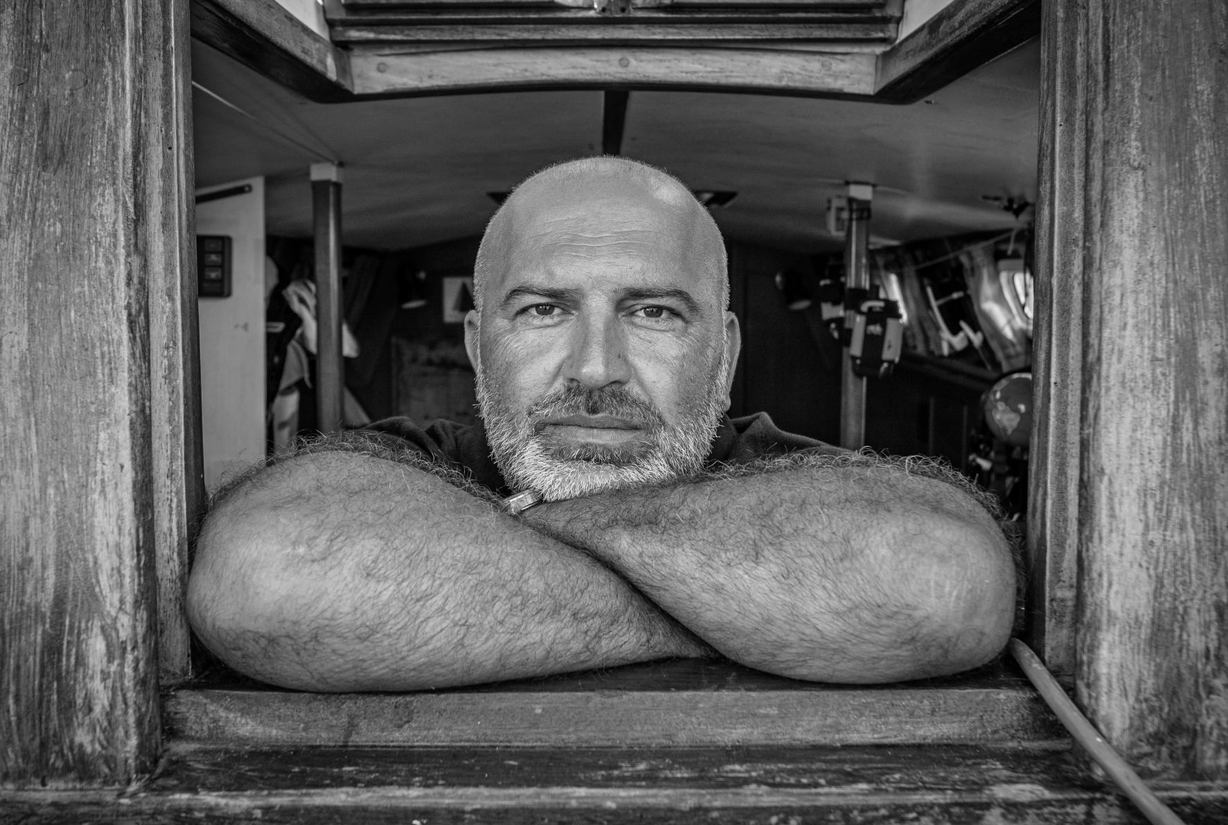 Skipper barbu accoudé au hublot en bois d’un voilier, portrait noir et blanc, regard intense, lumière contrastée