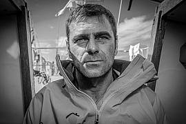 Portrait noir et blanc de Damien Guillou en veste de quart, regard déterminé dans la cabine de son voilier de course.