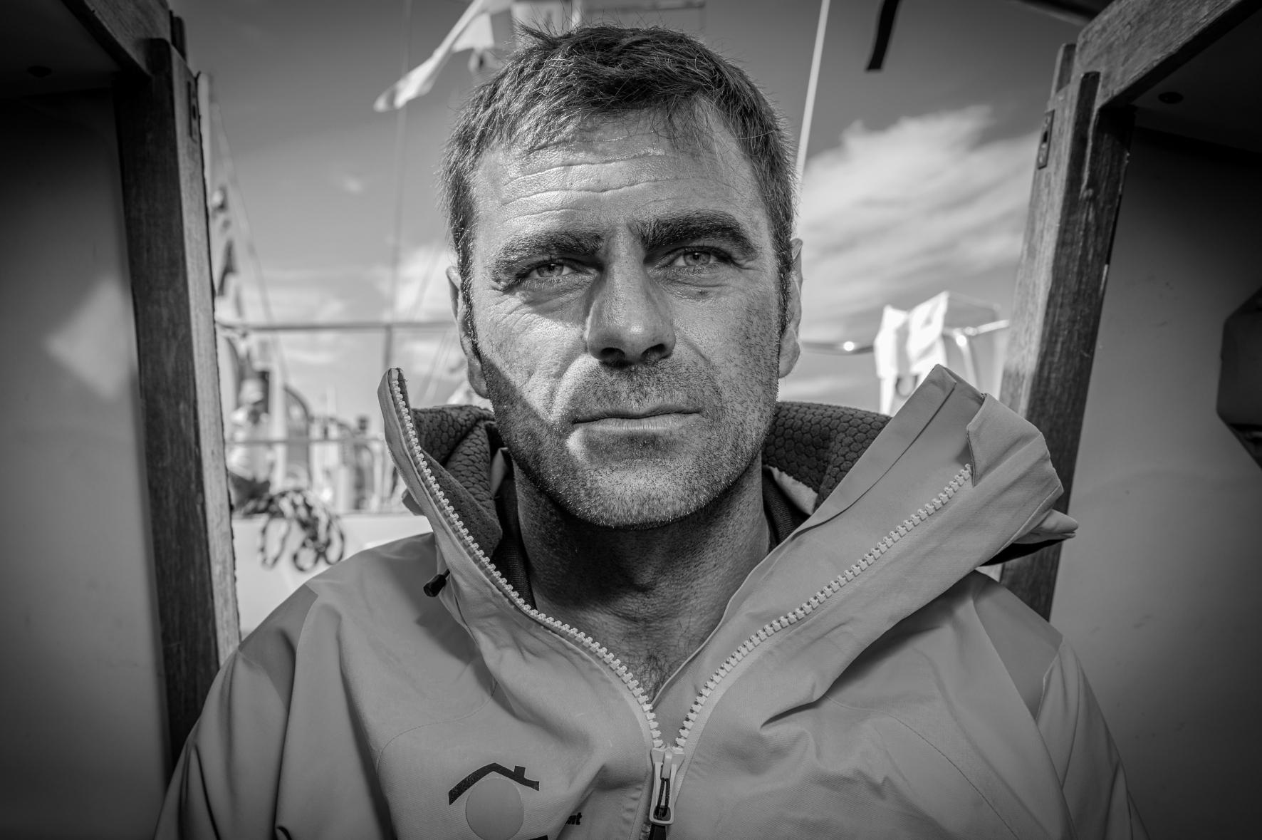 Portrait noir et blanc de Damien Guillou en veste de quart, regard déterminé dans la cabine de son voilier de course.