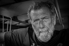 Portrait en noir et blanc d’un skipper barbu au regard intense dans le cockpit de son voilier de course.