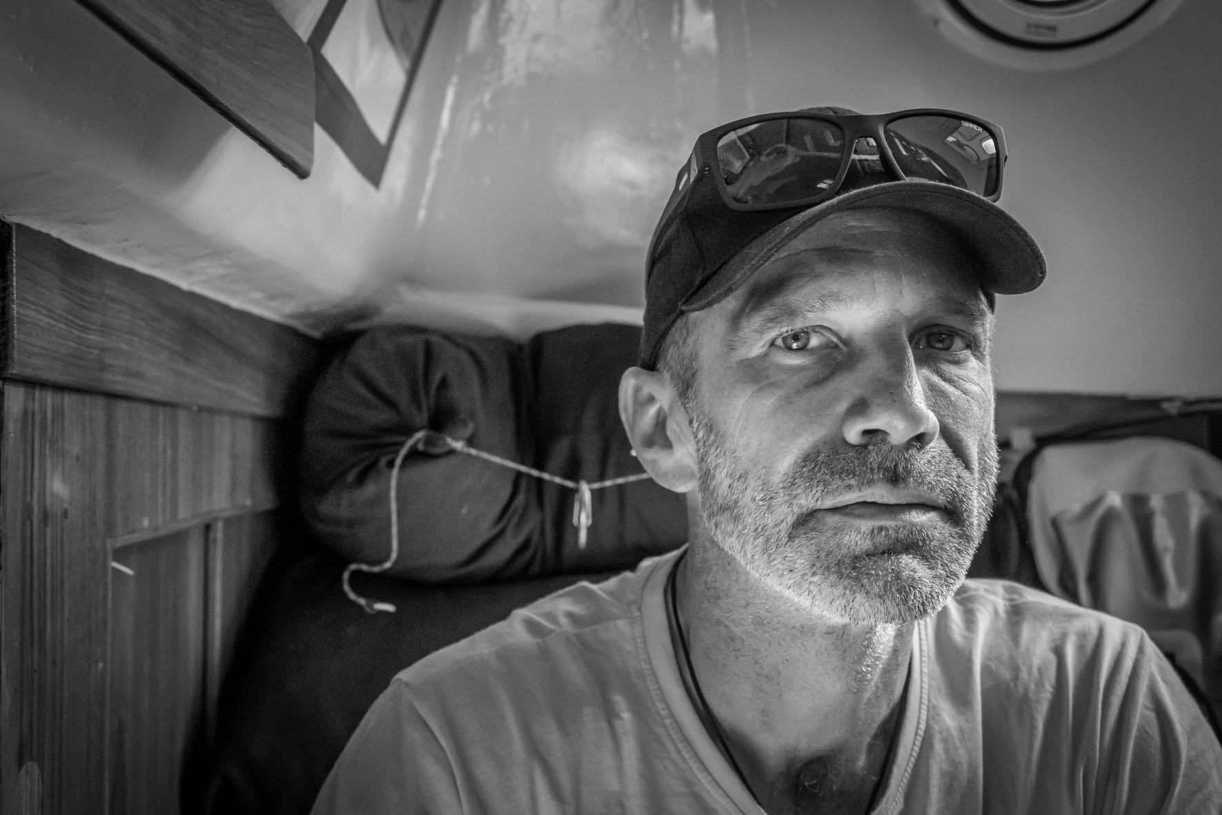 Portrait noir et blanc de Michael Guggenberger avec casquette et lunettes dans la cabine d’un voilier de course.