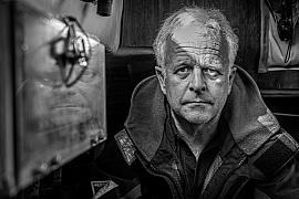 Portrait noir et blanc du skipper Jeremy Bagshaw à bord, regard intense et reflet sur vitre de cabine.