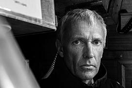 Portrait serré en noir et blanc de Guy Waites, regard intense, éclairage contrasté dans la cabine d’un voilier.