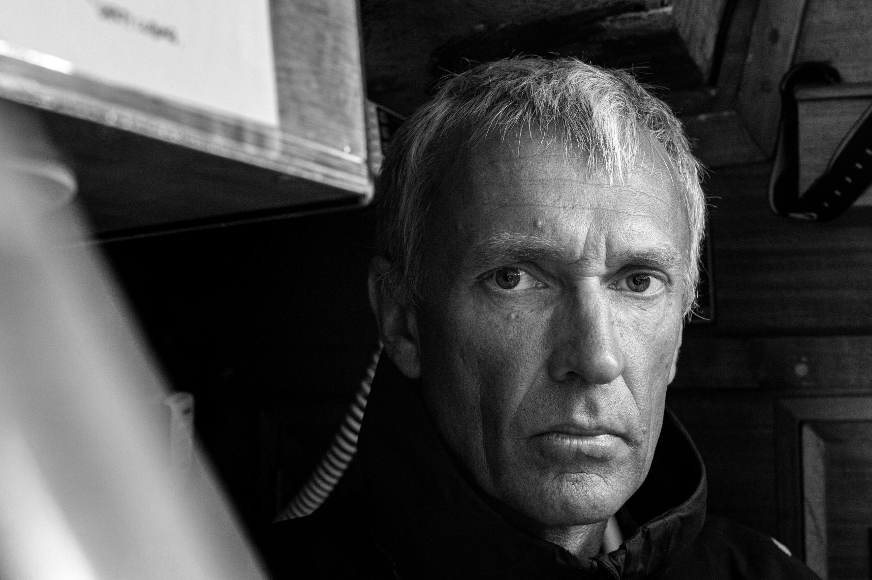 Portrait serré en noir et blanc de Guy Waites, regard intense, éclairage contrasté dans la cabine d’un voilier.