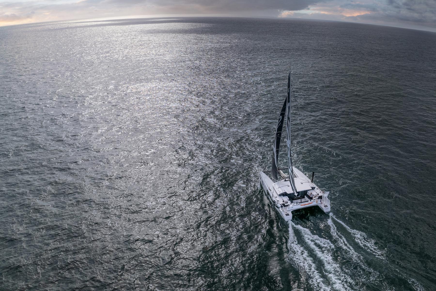 Vue aérienne d’un catamaran blanc sur mer sombre scintillante, ciel nuageux breton, éclairage naturel dramatique.