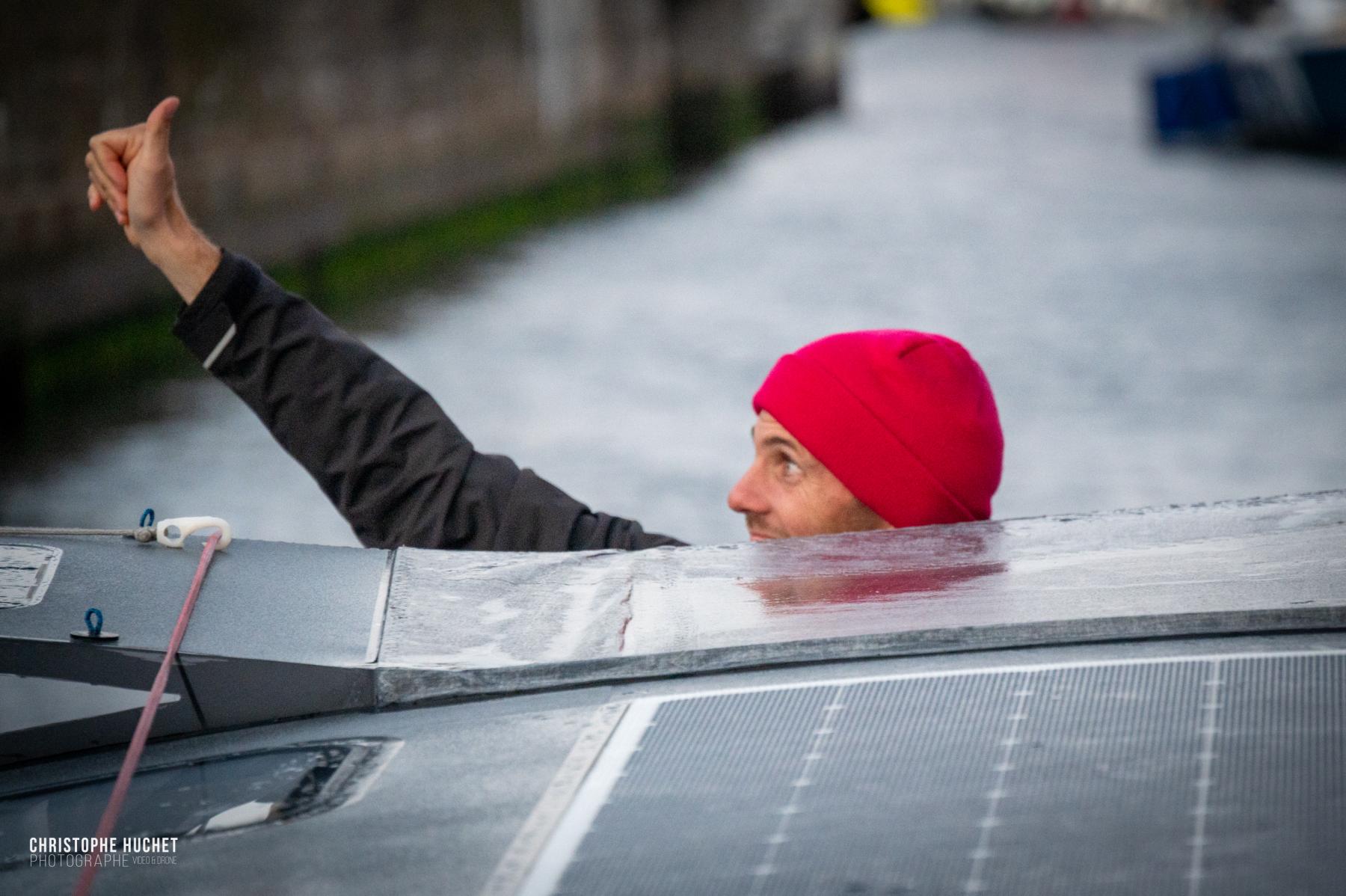 Skipper en bonnet rouge faisant un pouce levé depuis le pont de l’Imoca Lazare aux écluses de Saint-Malo.