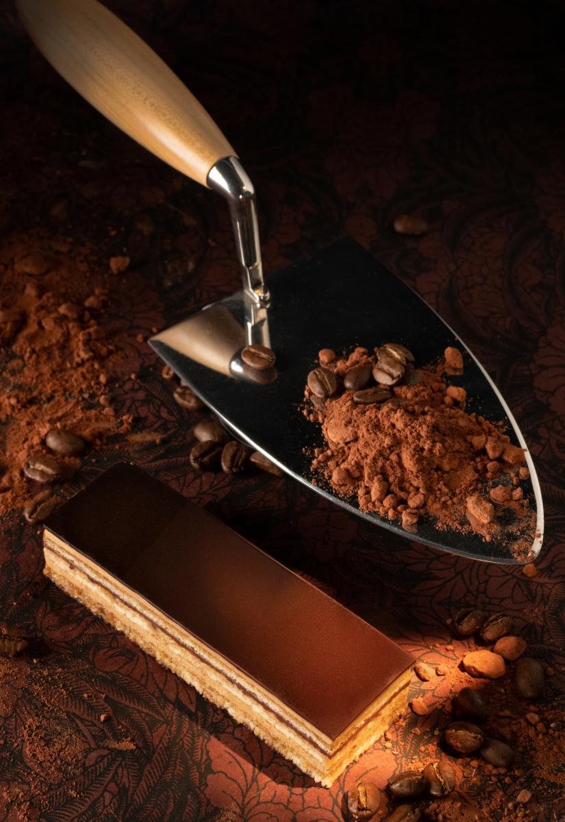 Gâteau opéra à côté d’une truelle d’acier chargée de poudre de cacao et grains de café, lumière contrastée