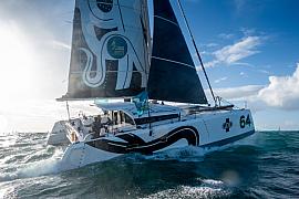 Catamaran blanc GDD fendant les vagues au large de Saint-Malo, voiles déployées sous un ciel marin lumineux.