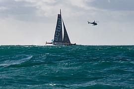 Trimaran Ultim Actual navigant sur une mer agitée sous un hélicoptère lors du départ de la course à Saint-Malo.
