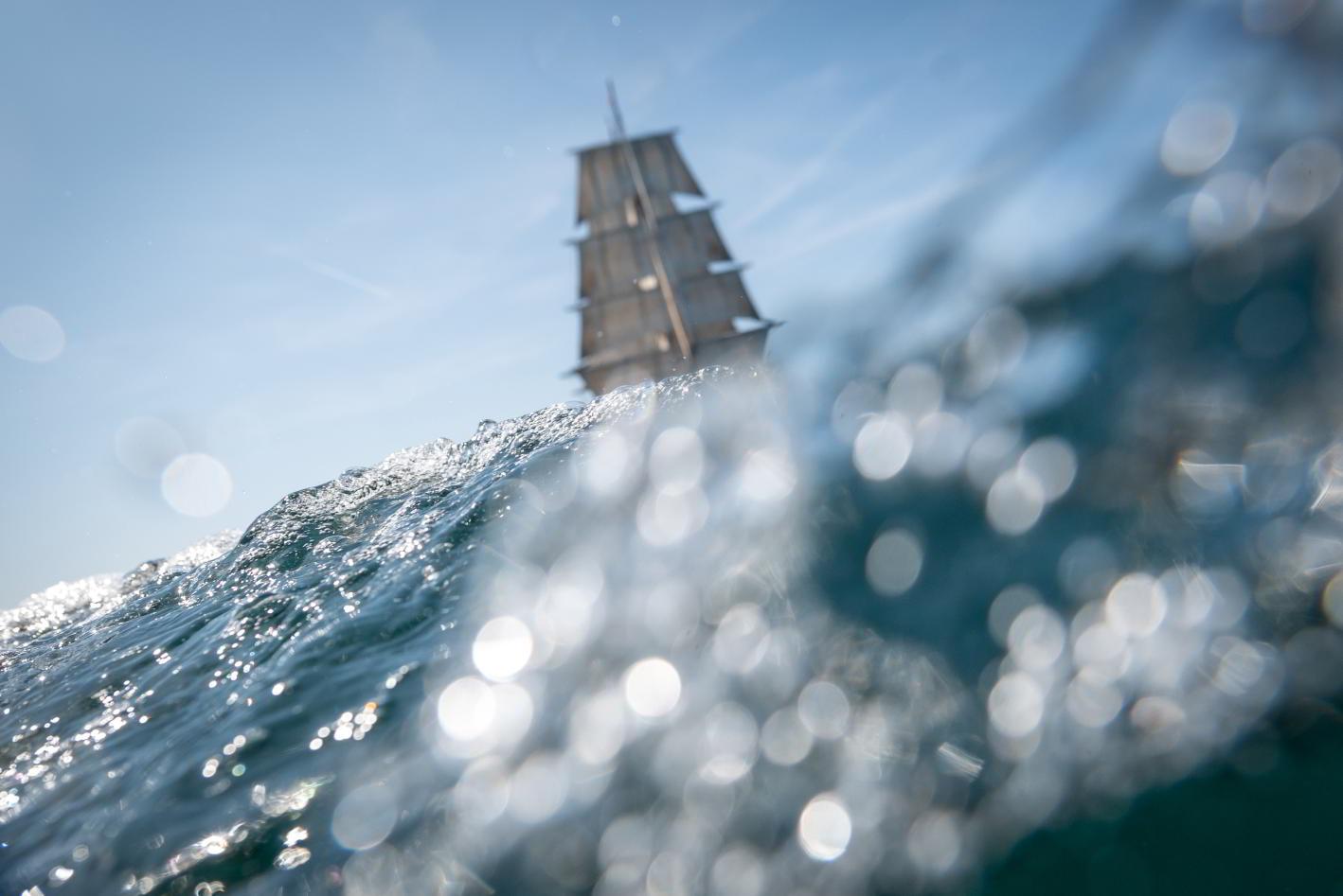 Vieux gréement à trois mâts vu au ras de l’eau à travers l’écume d’une vague sous un soleil éclatant.