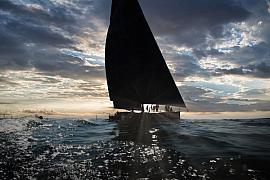 Silhouette de l’Imoca Lazare sur mer agitée, soleil perçant les nuages derrière la voile, vagues à l’avant-plan.