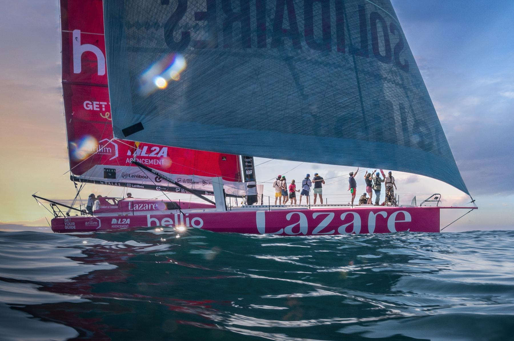 Voilier de course Imoca Lazare rose sur l’eau au coucher du soleil avec équipage sur le pont et voiles déployées.