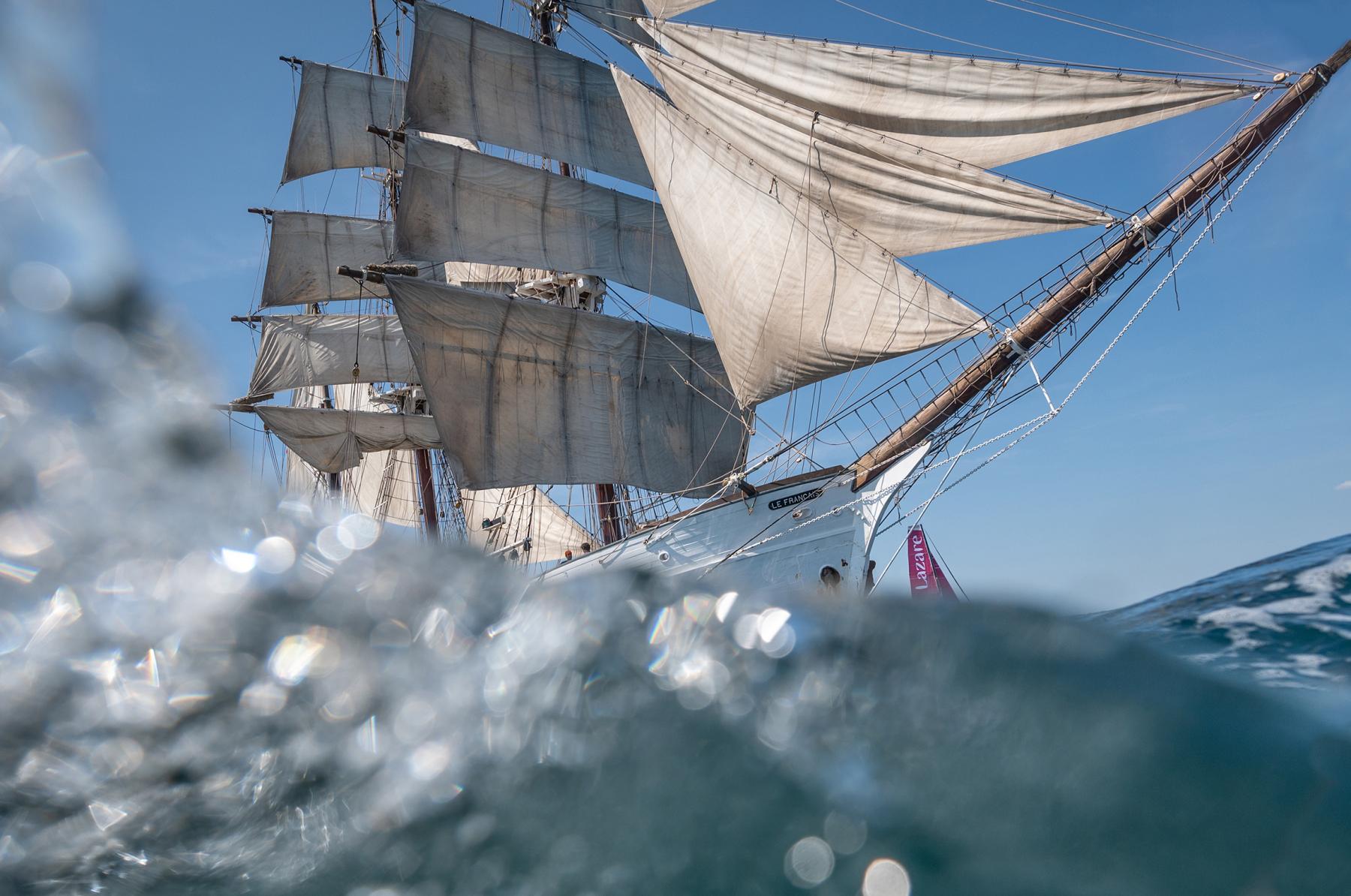 Trois-mâts Le Français sous voiles derrière une vague d’écume au premier plan, Imoca Lazare en arrière-plan.