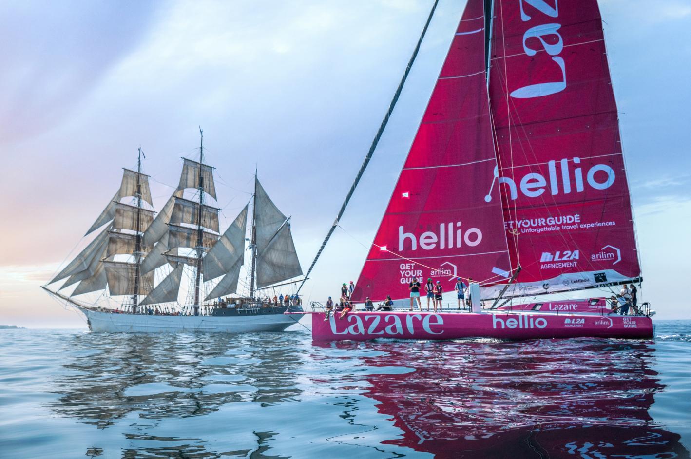 IMOCA rose Lazare et vieux gréement à trois mâts naviguant côte à côte en mer calme sous une lumière douce.