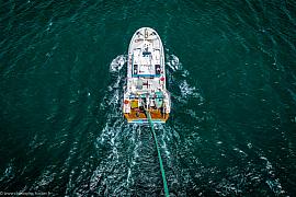 Vue drone d’un chalutier naviguant sur une mer emeraude profonde avec un sillage d’écume blanche et un chalut en cours de déploiement.