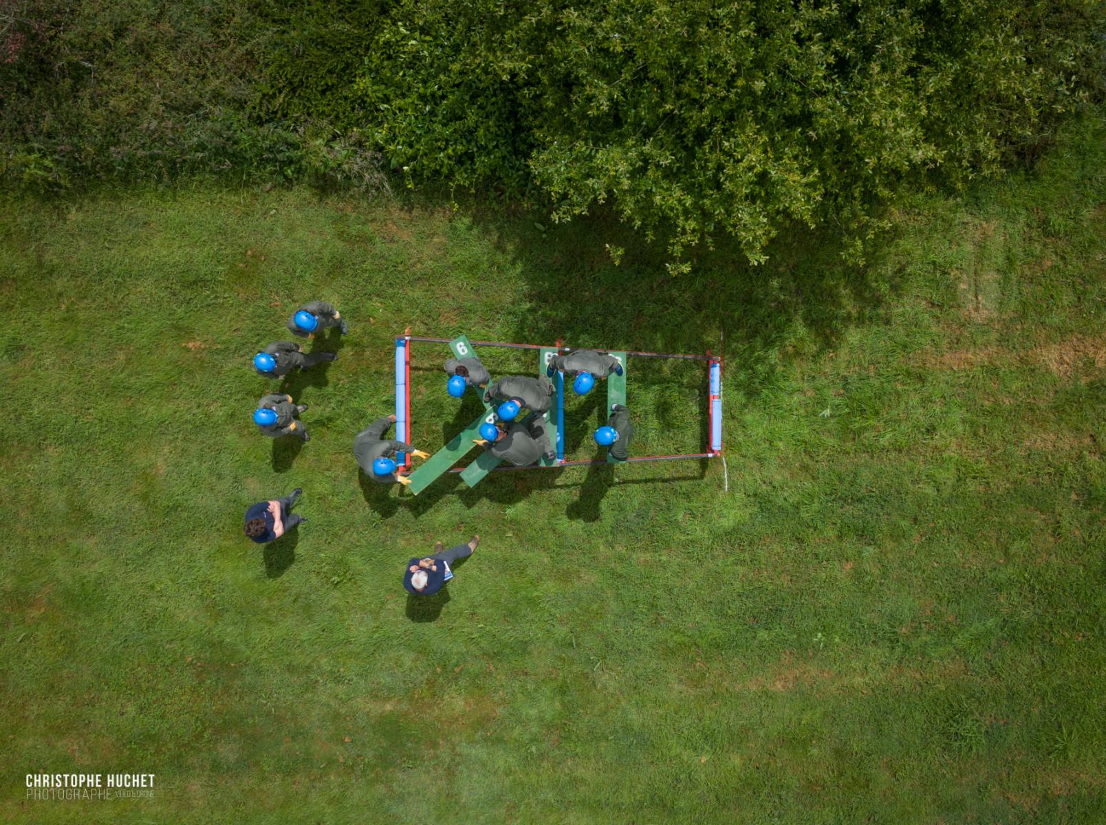 Vue drone zénitale de stagiaires en casques bleus déplaçant des madriers sur herbe lors d’un atelier leadership 
