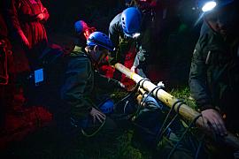 Équipe casquée avec lampes frontales manœuvrant un brancard de fortune lors d’un exercice nocturne en forêt.